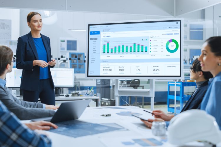 Modern Industrial Factory Meeting: Confident Female Engineer Uses Interactive Whiteboard, Makes Report to a Group of Engineers, Managers Talks and Shows Statistics, Growth and Analysis Information