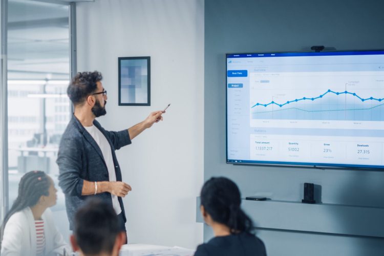 Diverse Modern Office: Motivated Businessman Leads Business Meeting with Managers, Talks, uses Presentation TV with Statistics, Chart Growth, Big Data. Digital Entrepreneurs Work on e-Commerce Project