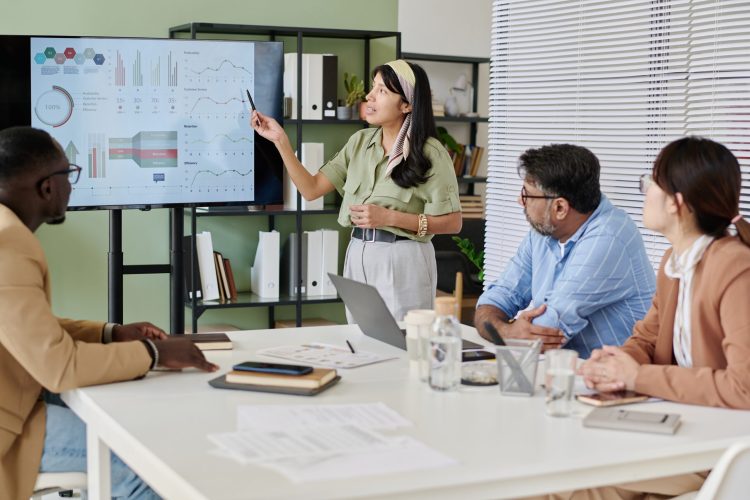 Group of colleagues discussing business data in meeting room with charts on screen and various documents on table. Presenter explaining graphs while others listening attentively